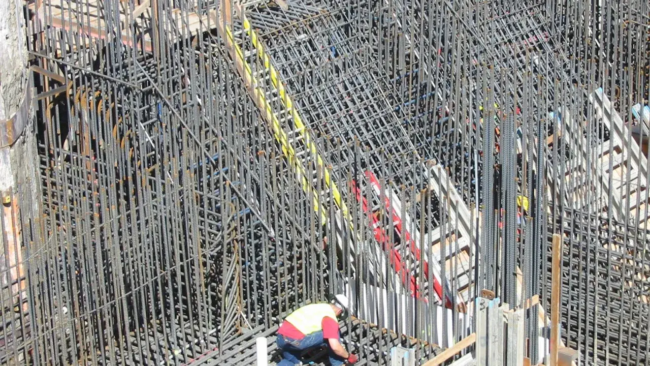 Operaio lavora su un'impalcatura di ferro per il cemento armato. La sua presenza suggerisce la cura necessaria per garantire la durata del cemento armato.