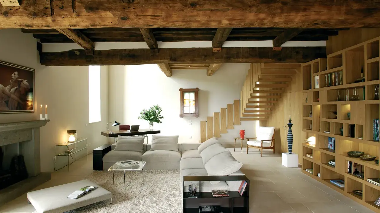 Soggiorno moderno con travi a vista, un camino in pietra, divani chiari, libreria in legno e scala a vista. Le travi a vista pro e contro sono evidenti nell'estetica rustica e nella potenziale manutenzione.