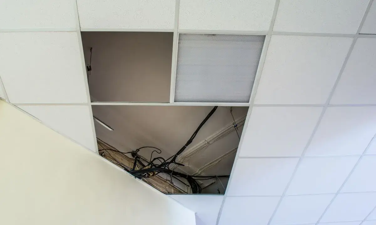 Controsoffitto in fibra con pannelli rimossi che espongono cavi e tubi. Un pannello con griglia luminosa è visibile.