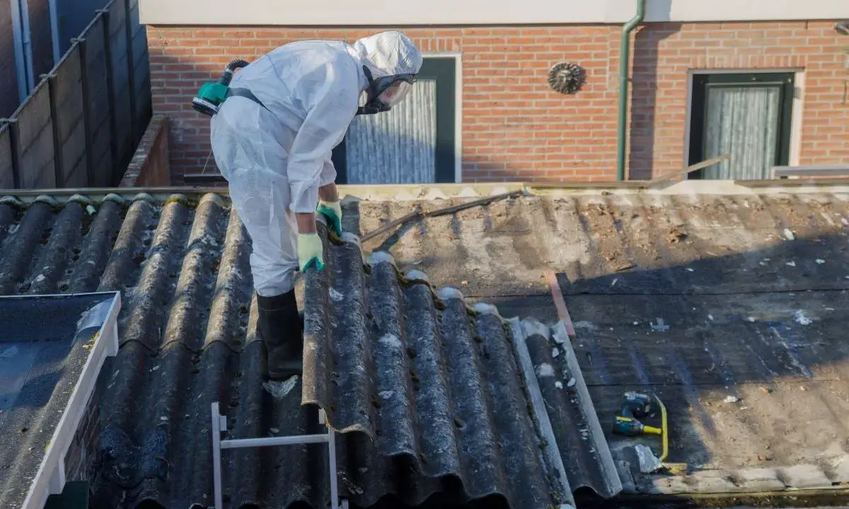 Operaio in tuta protettiva rimuove eternit da tetto di un edificio in linea.