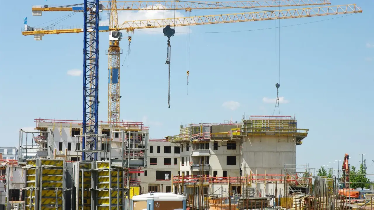 Grandi gru gialle e blu dominano un cantiere edile con edifici in costruzione. Sembra che il progetto abbia subito mille proroghe, con materiali sparsi e strutture incomplete.