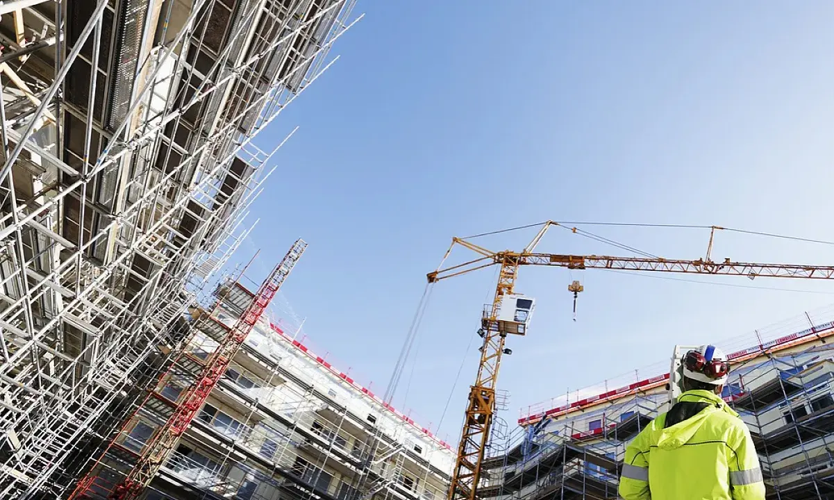 Operaio con giubbotto alta visibilità osserva un cantiere edile con gru e impalcature. Capire cassa edile come funziona è fondamentale per la sicurezza.