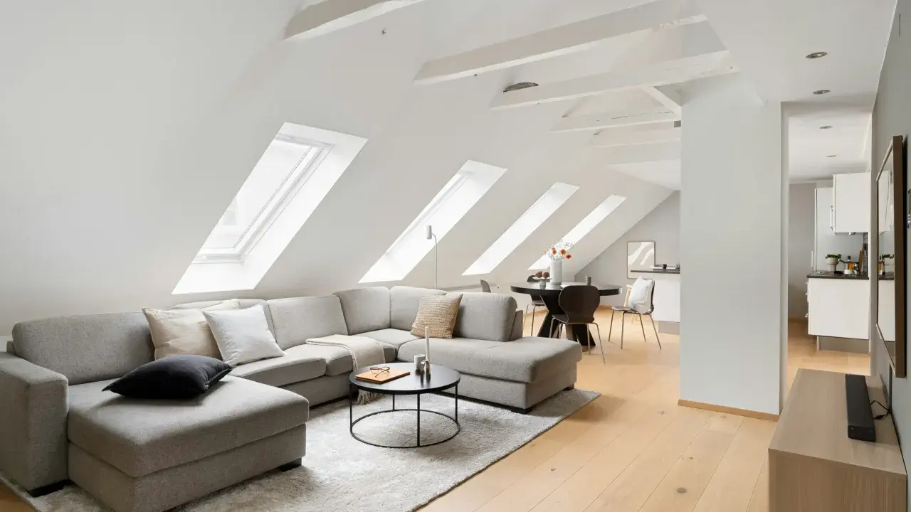 Soggiorno mansardato con divano angolare grigio, tavolino nero e travi in legno a vista sul soffitto inclinato.