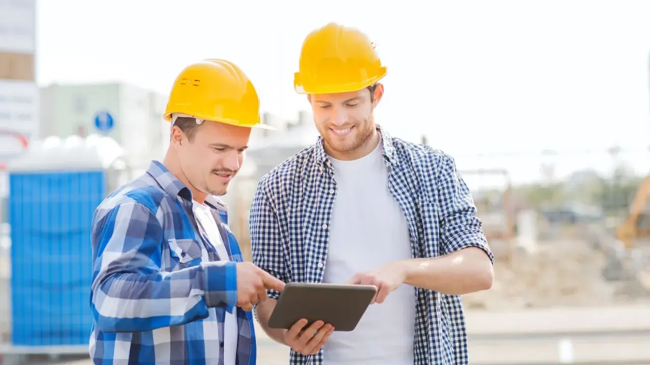 Due operai con caschi gialli esaminano un tablet, forse per un verbale di sopralluogo cantiere. Sullo sfondo, un cantiere con bagni chimici.