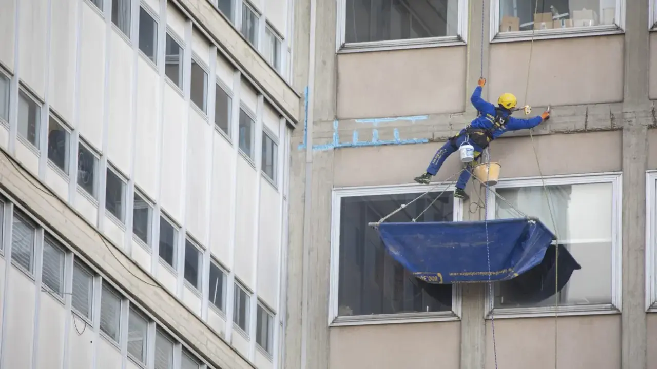 Operaio in corda esegue consolidamenti strutturali su un edificio, con attrezzature di sicurezza e secchi.