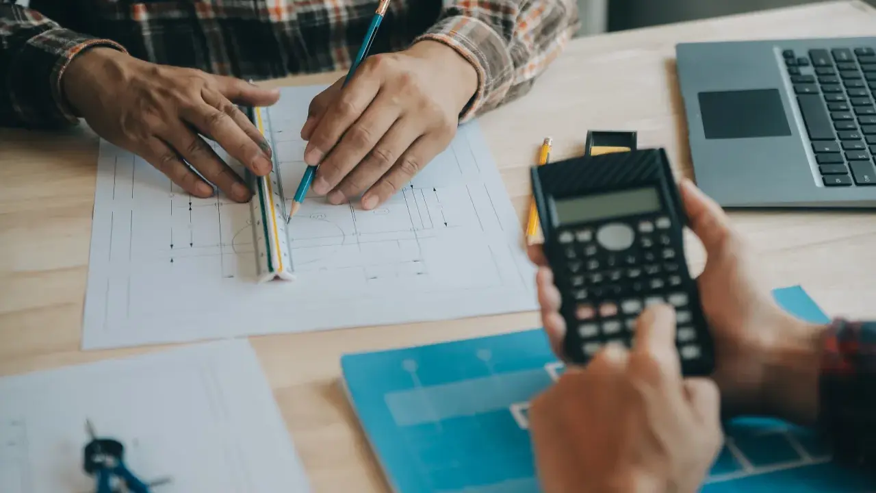 Due mani lavorano su progetti, una usa una calcolatrice, l'altra una matita e un righello, per definire i requisiti igienico sanitari.