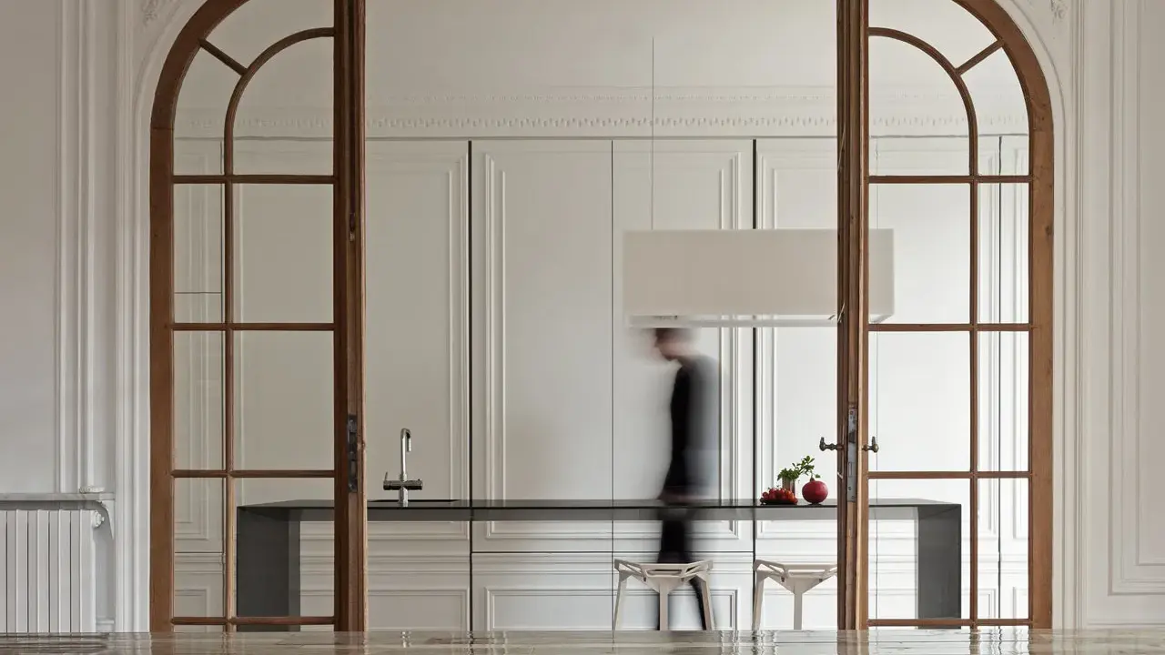 Archi interni casa con porte in legno che incorniciano una cucina moderna. Una figura sfocata attraversa lo spazio.
