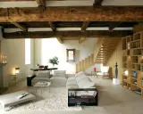 Soggiorno moderno con travi a vista, un camino in pietra, divani chiari, libreria in legno e scala a vista. Le travi a vista pro e contro sono evidenti nell'estetica rustica e nella potenziale manutenzione.