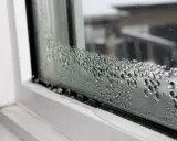 Goccioline d'acqua formano una **condensa interstiziale** sul vetro di una finestra, con un telaio bianco visibile.