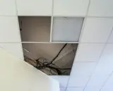 Controsoffitto in fibra con pannelli rimossi che espongono cavi e tubi. Un pannello con griglia luminosa è visibile.
