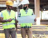Due operai con caschi e giubbotti ad alta visibilità esaminano un progetto. Uno, il responsabile dei lavori, indica un dettaglio.
