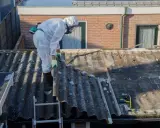 Operaio in tuta protettiva rimuove eternit da tetto di un edificio in linea.