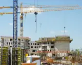 Grandi gru gialle e blu dominano un cantiere edile con edifici in costruzione. Sembra che il progetto abbia subito mille proroghe, con materiali sparsi e strutture incomplete.