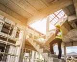 Ingegnere con caschetto giallo e giubbotto ad alta visibilità, con progetti in mano, osserva la **soletta edilizia** in cemento grezzo di un cantiere in costruzione.