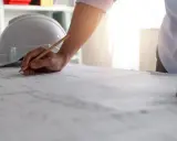 Un ingegnere esamina i disegni per lo stato avanzamento lavori privati, con un casco e un metro sul tavolo.