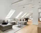 Soggiorno mansardato con divano angolare grigio, tavolino nero e travi in legno a vista sul soffitto inclinato.