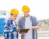 Due operai con caschi gialli esaminano un tablet, forse per un verbale di sopralluogo cantiere. Sullo sfondo, un cantiere con bagni chimici.