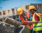 Due operai con caschi gialli e giubbotti ad alta visibilità discutono un progetto di **construction management** su un cantiere.