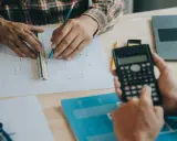Due mani lavorano su progetti, una usa una calcolatrice, l'altra una matita e un righello, per definire i requisiti igienico sanitari.