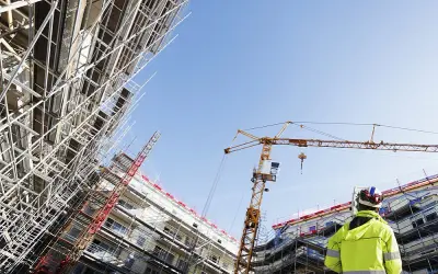 Operaio con giubbotto alta visibilità osserva un cantiere edile con gru e impalcature. Capire cassa edile come funziona è fondamentale per la sicurezza.