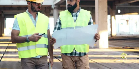 Due operai con caschi e giubbotti ad alta visibilità esaminano un progetto. Uno, il responsabile dei lavori, indica un dettaglio.