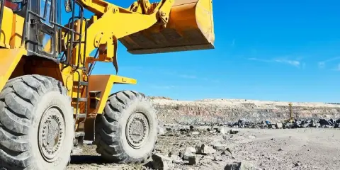 Pale giallo escavatore solleva un carico di terre e rocce da scavo in una cava sotto un cielo azzurro.