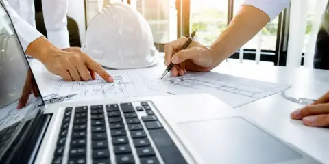 Ingegneri esaminano un **elenco SOA** e progetti su un tavolo con un casco bianco e un laptop.