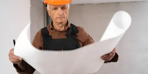 Un esperto capomastro, con casco arancione, studia attentamente un progetto, dimostrando il vero significato di guida e competenza in cantiere.