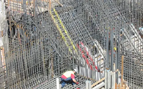 Operaio lavora su un'impalcatura di ferro per il cemento armato. La sua presenza suggerisce la cura necessaria per garantire la durata del cemento armato.
