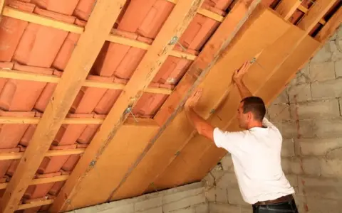 Operaio posiziona pannelli per isolamento soffitto interno, tra travi in legno e tegole.