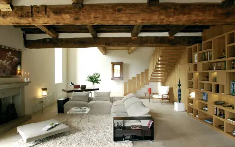 Soggiorno moderno con travi a vista, un camino in pietra, divani chiari, libreria in legno e scala a vista. Le travi a vista pro e contro sono evidenti nell'estetica rustica e nella potenziale manutenzione.