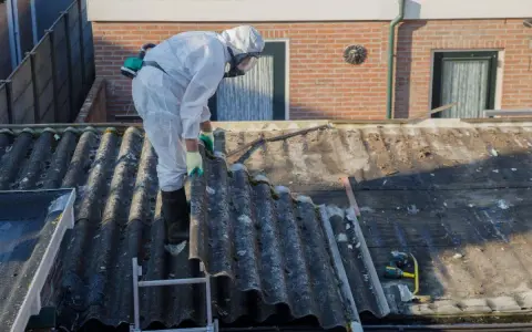 Operaio in tuta protettiva rimuove eternit da tetto di un edificio in linea.