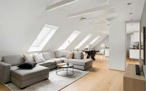 Soggiorno mansardato con divano angolare grigio, tavolino nero e travi in legno a vista sul soffitto inclinato.