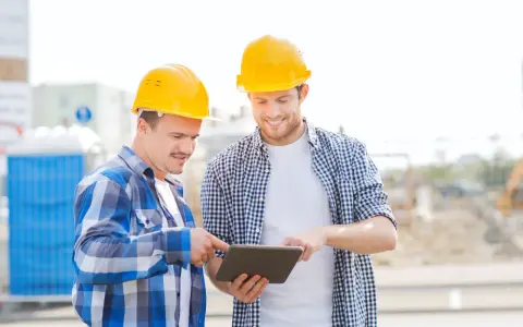 Due operai con caschi gialli esaminano un tablet, forse per un verbale di sopralluogo cantiere. Sullo sfondo, un cantiere con bagni chimici.