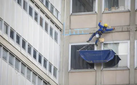 Operaio in corda esegue consolidamenti strutturali su un edificio, con attrezzature di sicurezza e secchi.