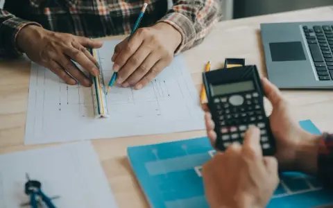 Due mani lavorano su progetti, una usa una calcolatrice, l'altra una matita e un righello, per definire i requisiti igienico sanitari.