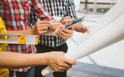 Team di operai esamina progetto su tablet, livella e rotoli di carta. Aggiornamento sal stato avanzamento lavori.
