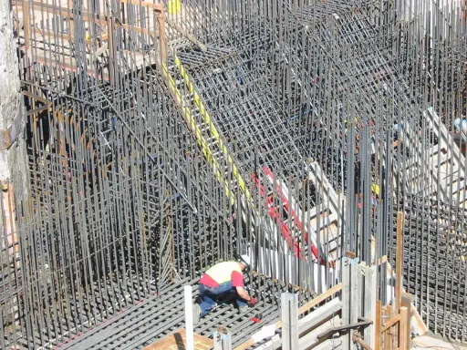 Operaio lavora su un'impalcatura di ferro per il cemento armato. La sua presenza suggerisce la cura necessaria per garantire la durata del cemento armato.