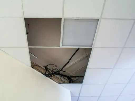 Controsoffitto in fibra con pannelli rimossi che espongono cavi e tubi. Un pannello con griglia luminosa è visibile.