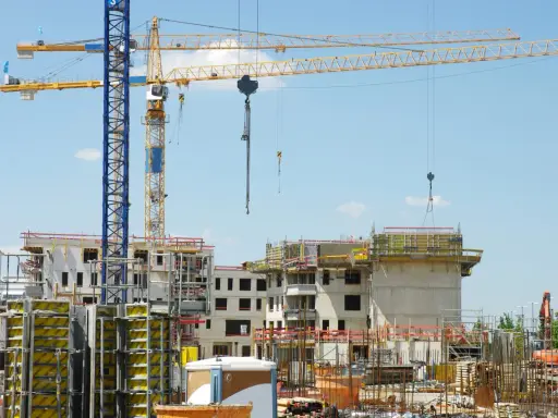 Grandi gru gialle e blu dominano un cantiere edile con edifici in costruzione. Sembra che il progetto abbia subito mille proroghe, con materiali sparsi e strutture incomplete.