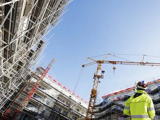 Operaio con giubbotto alta visibilità osserva un cantiere edile con gru e impalcature. Capire cassa edile come funziona è fondamentale per la sicurezza.