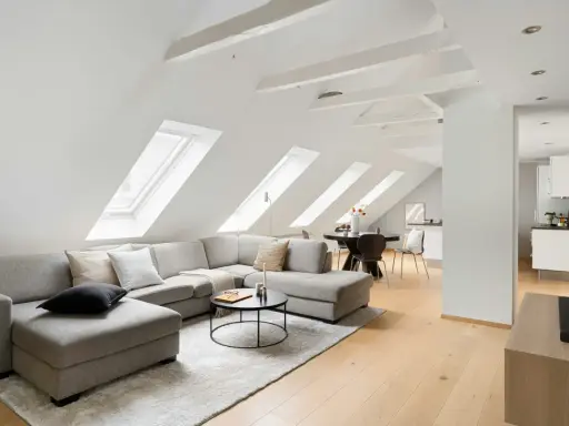 Soggiorno mansardato con divano angolare grigio, tavolino nero e travi in legno a vista sul soffitto inclinato.