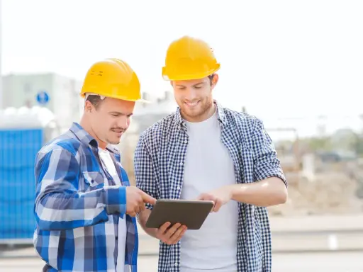 Due operai con caschi gialli esaminano un tablet, forse per un verbale di sopralluogo cantiere. Sullo sfondo, un cantiere con bagni chimici.