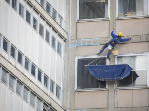 Operaio in corda esegue consolidamenti strutturali su un edificio, con attrezzature di sicurezza e secchi.