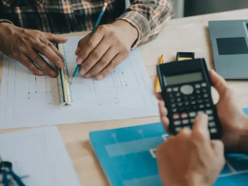 Due mani lavorano su progetti, una usa una calcolatrice, l'altra una matita e un righello, per definire i requisiti igienico sanitari.