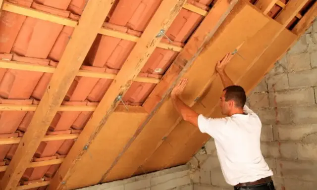 Operaio posiziona pannelli per isolamento soffitto interno, tra travi in legno e tegole.
