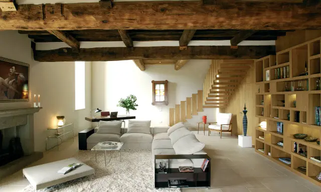 Soggiorno moderno con travi a vista, un camino in pietra, divani chiari, libreria in legno e scala a vista. Le travi a vista pro e contro sono evidenti nell'estetica rustica e nella potenziale manutenzione.