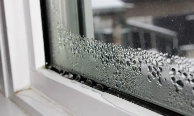 Goccioline d'acqua formano una **condensa interstiziale** sul vetro di una finestra, con un telaio bianco visibile.