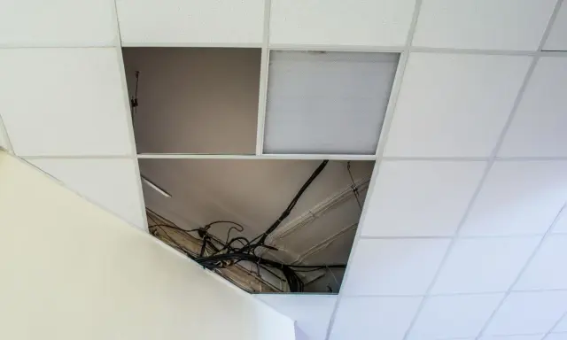Controsoffitto in fibra con pannelli rimossi che espongono cavi e tubi. Un pannello con griglia luminosa è visibile.