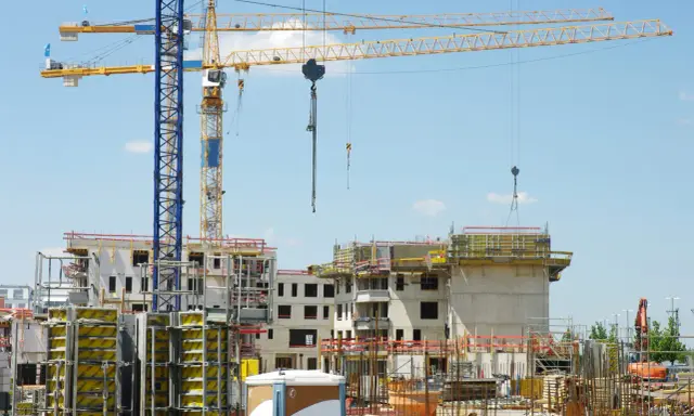 Grandi gru gialle e blu dominano un cantiere edile con edifici in costruzione. Sembra che il progetto abbia subito mille proroghe, con materiali sparsi e strutture incomplete.